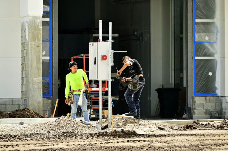 Muncitor într-o cască galbenă privește spre un utilaj de construcții sub soarele de vară